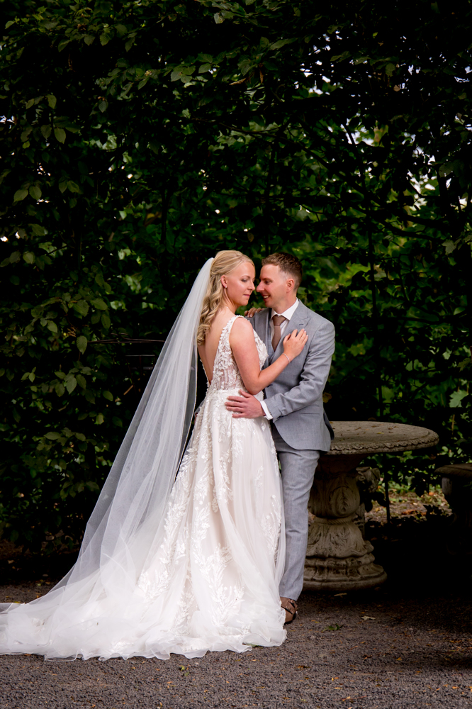 Real Bride Zohra & Jort – bruid en bruidegom omhelzen elkaar liefdevol in een groene tuinsetting; Zohra draagt een romantische A-lijn trouwjurk met verfijnde kanten details, open rug en lange sluier voor een elegante en tijdloze uitstraling.